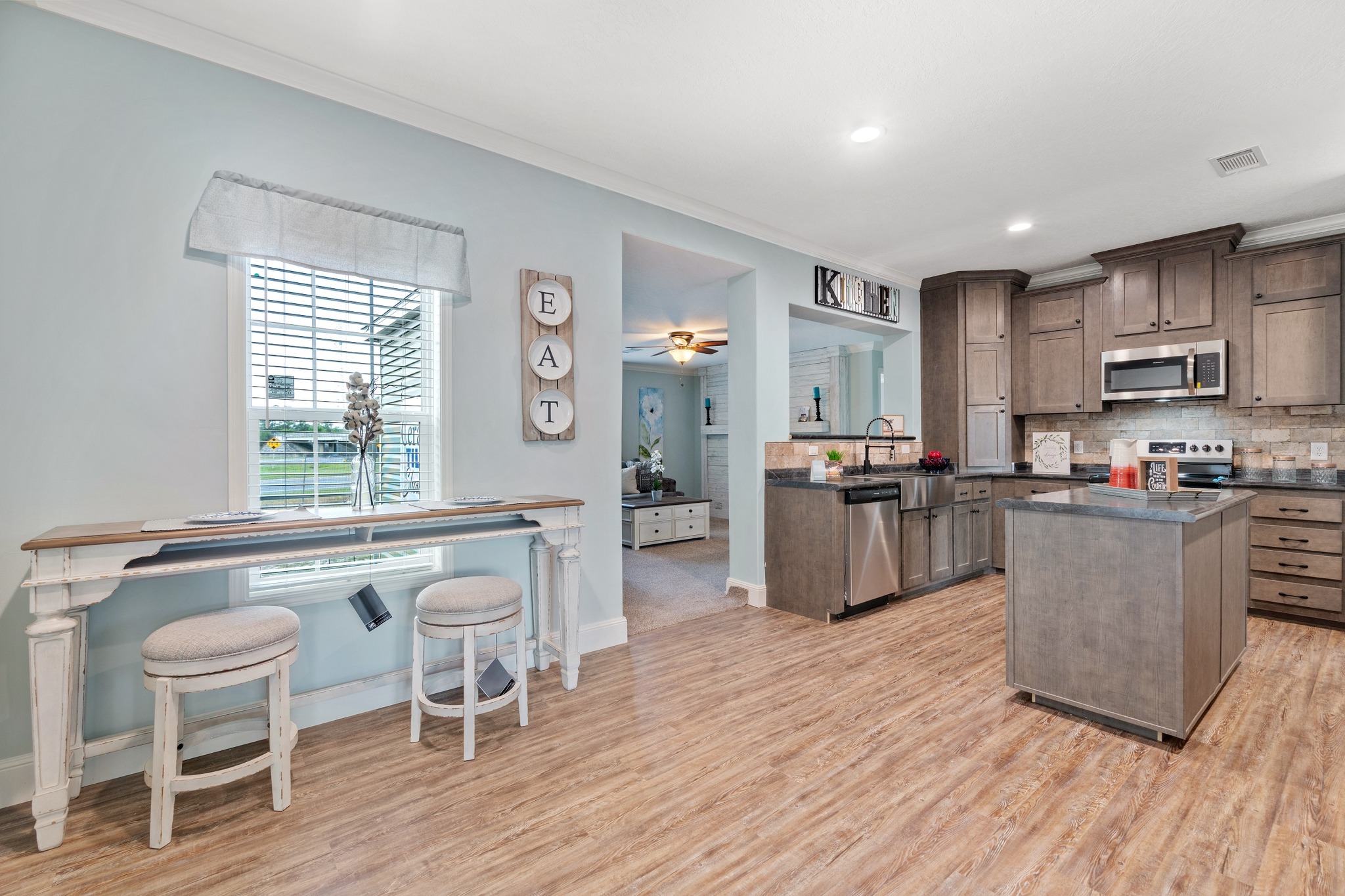Modern kitchen with wood cabinets and island, stainless steel appliances, and light blue walls. A small table and stools by the window add charm.