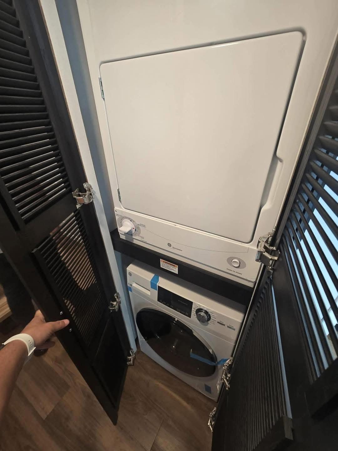 Stacked washer and dryer in a compact closet with black louvered doors, creating a neat and efficient laundry space. A hand partially opens a door.