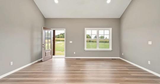 A spacious, empty room features gray walls, light wooden floors, and a white-trimmed window showing greenery. An open door leads to an outside view, conveying a calm, inviting atmosphere.