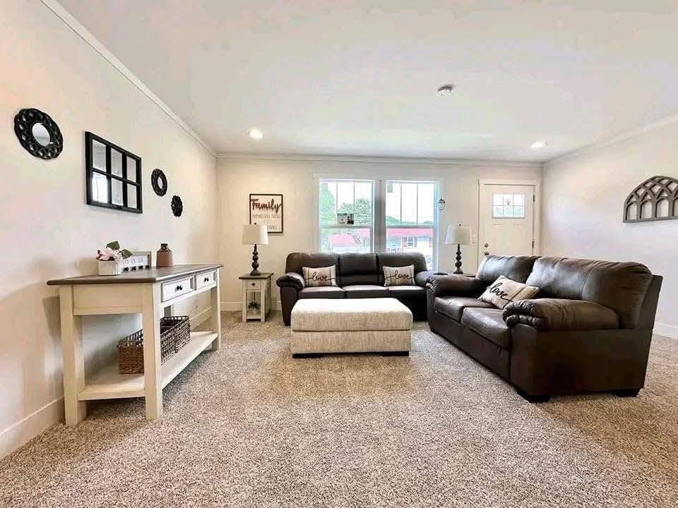 A cozy living room with beige walls features two dark brown sofas and a cream ottoman. Decor includes pillows with "Love," wall decor, and a white console table with baskets. Soft lighting provides a warm, inviting atmosphere.