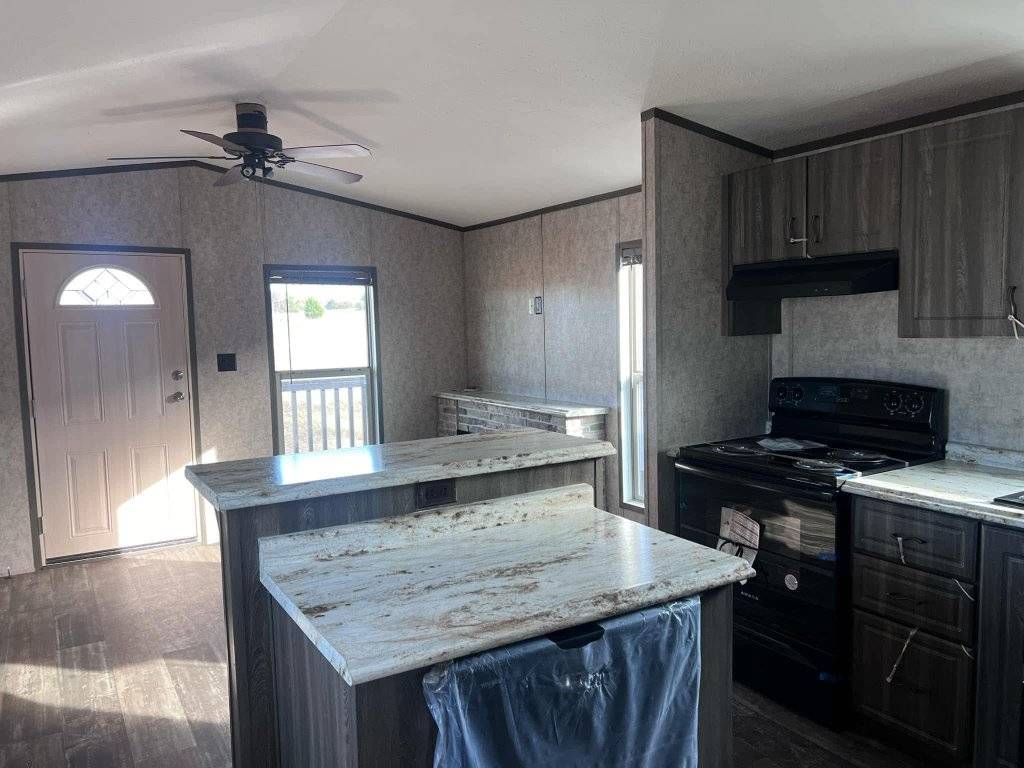 A modern kitchen with dark wood cabinets, an island with a marble countertop, and a black stove. Sunlight filters through a window near the entrance, creating a bright and inviting atmosphere.