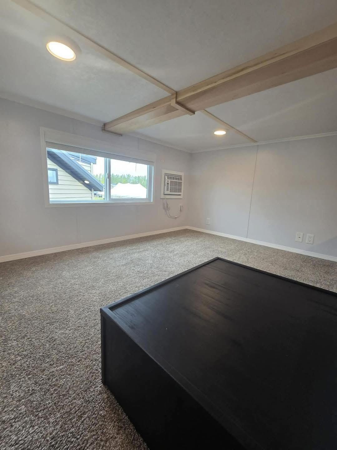 A spacious, carpeted room with gray walls, a large window, and ceiling lights. An air conditioning unit is mounted on the wall, and a black table is in the corner.