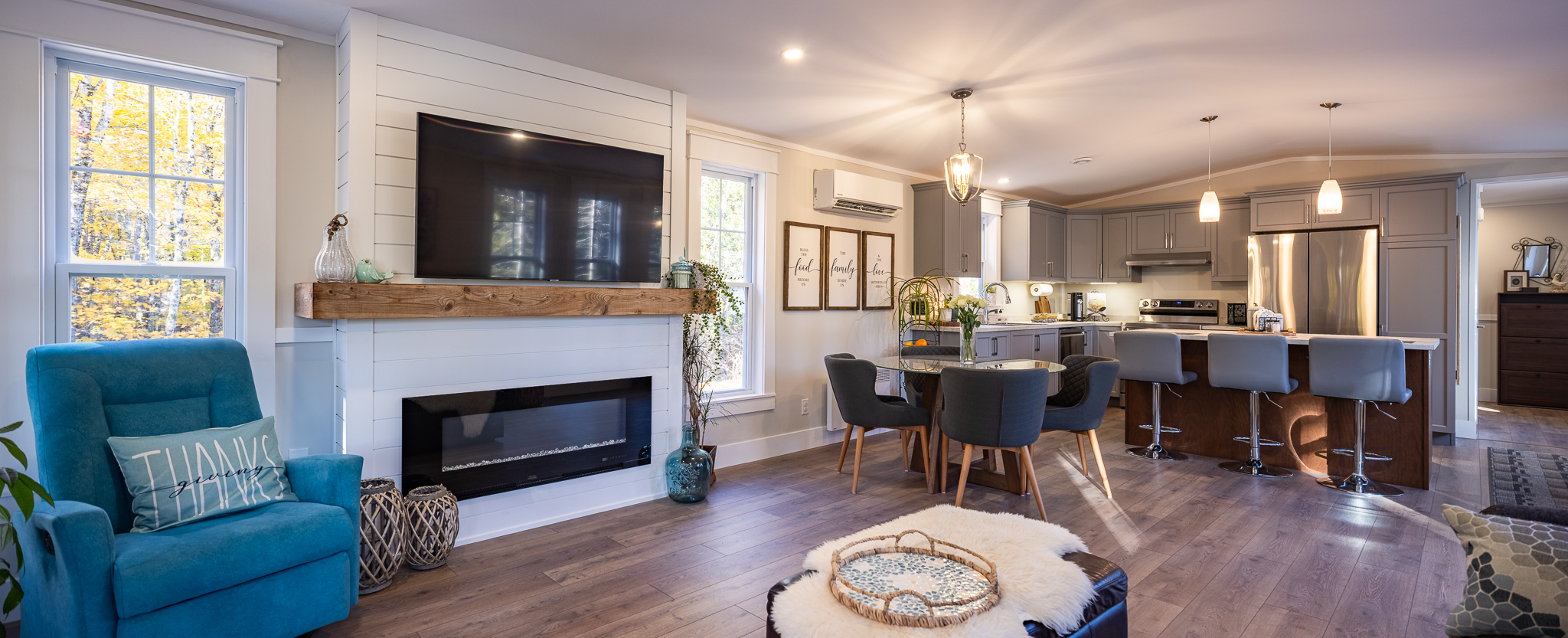 A cozy, modern open-concept living room and kitchen. Features a blue armchair, wall-mounted TV, fireplace, dining table, and a sleek kitchen with gray cabinets.