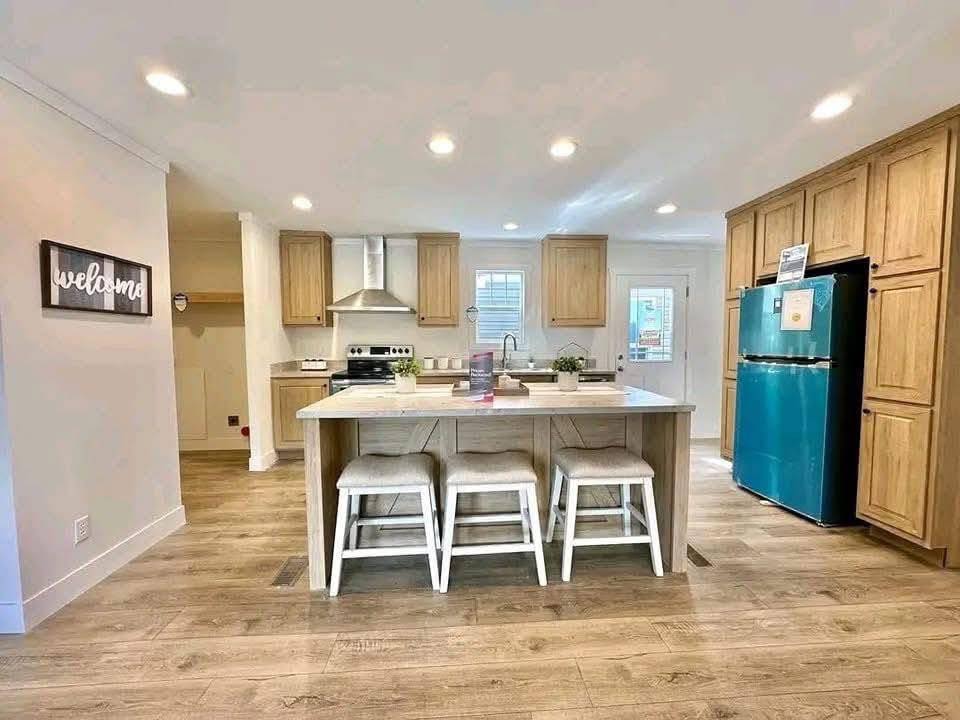 Bright kitchen with a central island featuring three stools, wood cabinets, a teal fridge, and a welcoming sign. Warm, inviting, and modern ambiance.