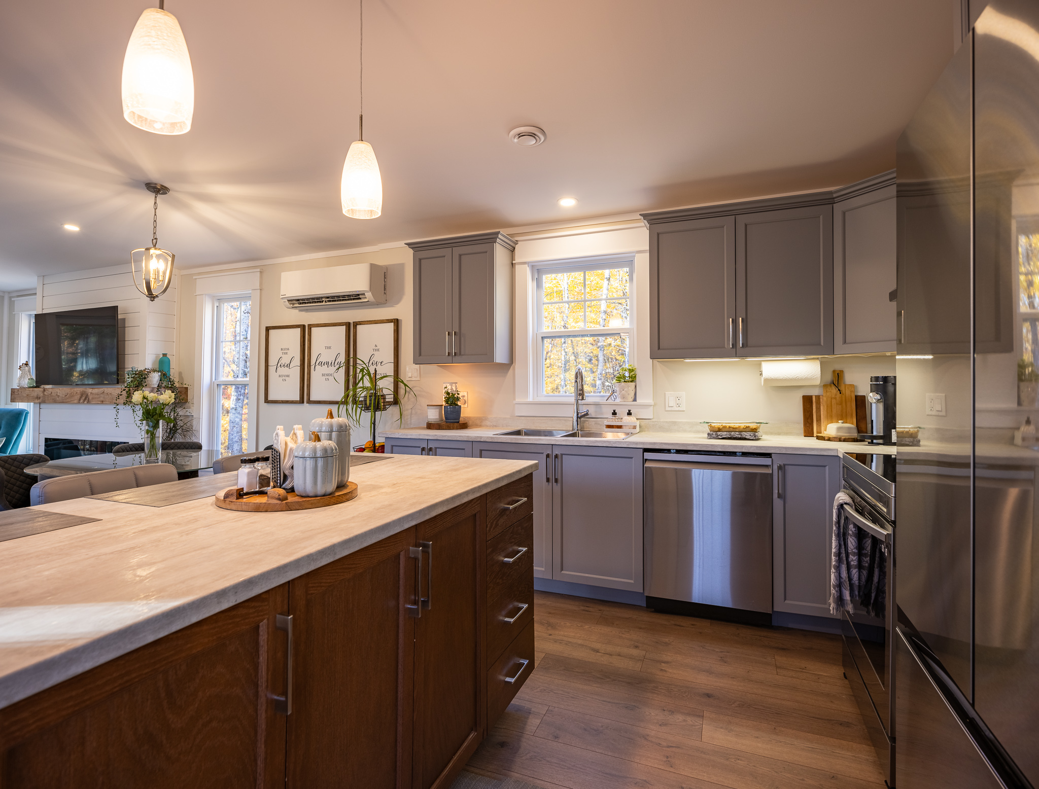 A modern kitchen with gray cabinets, wood flooring, and stainless steel appliances. A large island features decorative jars and pendant lighting.