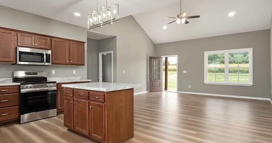 Spacious kitchen and living area with vaulted ceiling. Features stainless appliances, wood cabinets, island, open door, large window, and smooth flooring.