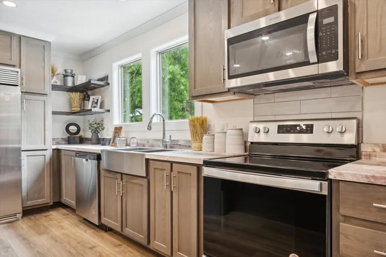 Modern kitchen with stainless steel appliances, light wood cabinets, and a farmhouse sink. Bright natural light from two windows creates a warm, inviting atmosphere.