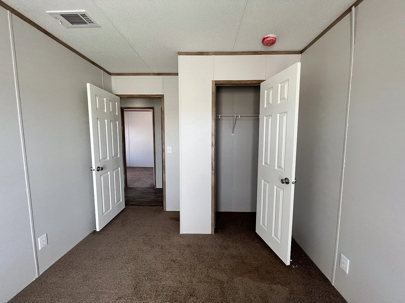 Empty room with beige walls and brown carpet. Two open doors reveal a closet and adjacent room. A red fire alarm is on the ceiling, conveying simplicity.