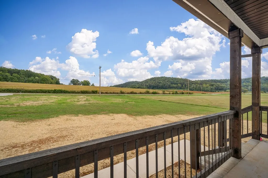View from a porch overlooking a vast, open field with a backdrop of distant hills and a bright blue sky dotted with fluffy clouds. The scene is serene and expansive.