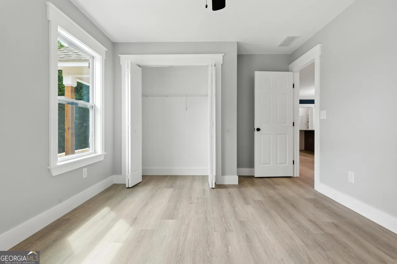 Bright, empty room with light wood flooring, gray walls, white trim, a double window on the left, open closet in the center, and a door on the right.