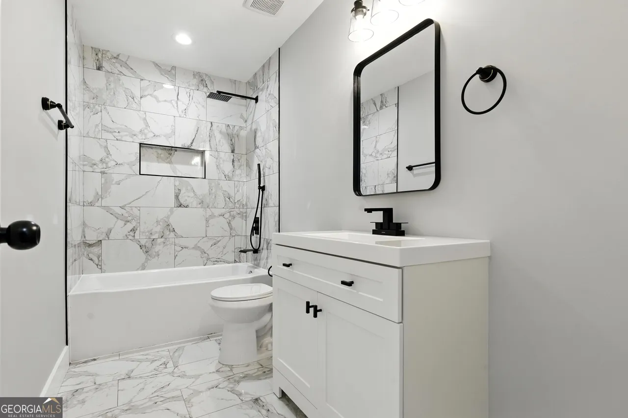 Modern bathroom with white marble tiles and a bathtub-shower combo. A black framed mirror and light fixtures contrast with white cabinetry, creating a sleek, clean look.