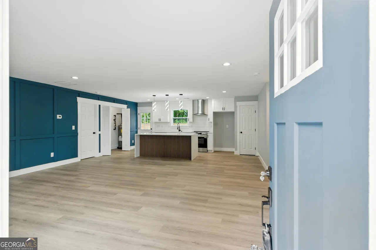 Open-concept living space with light wood flooring, a navy accent wall, and modern kitchen. Blue front door adds welcoming touch. Bright and airy.