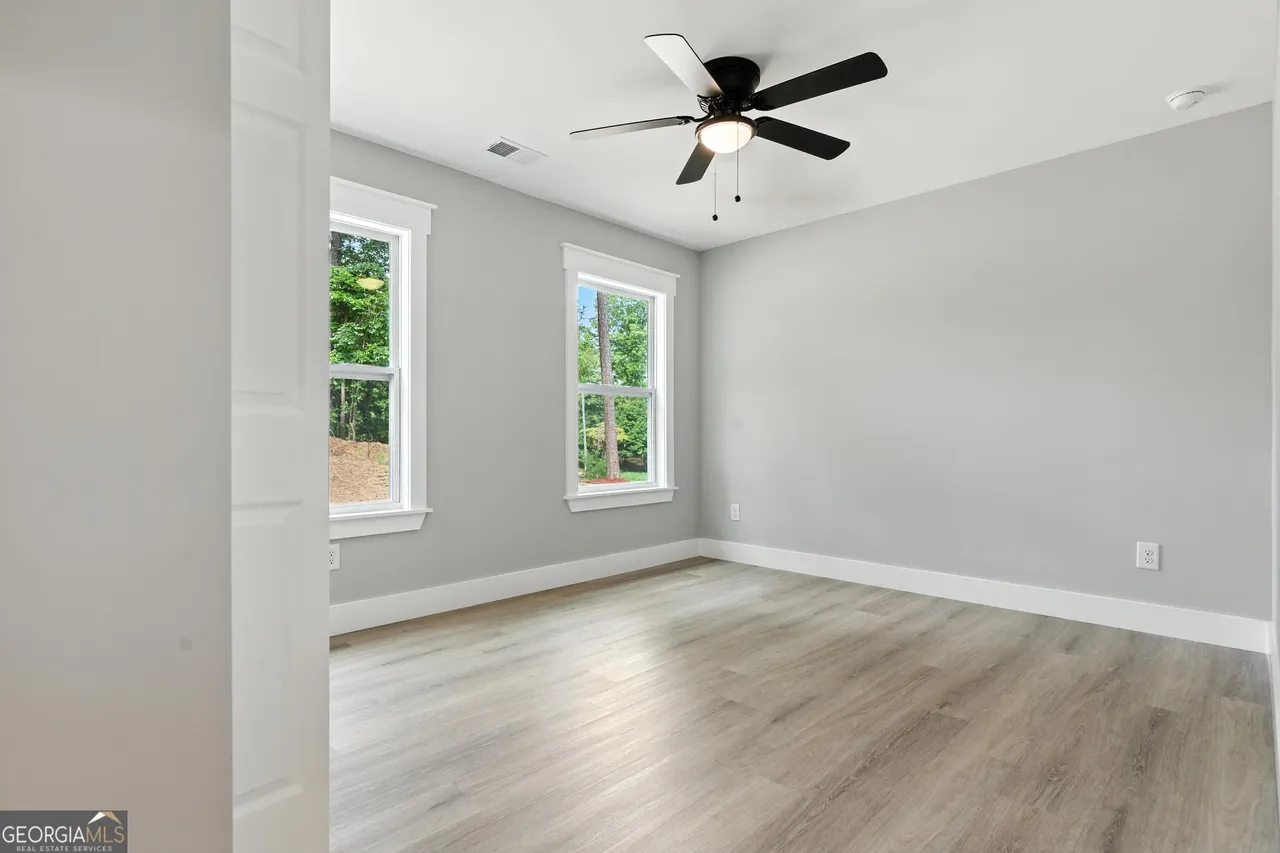 Minimalist room with light gray walls and wood flooring, featuring two large windows with a view of trees. A black ceiling fan hangs above.