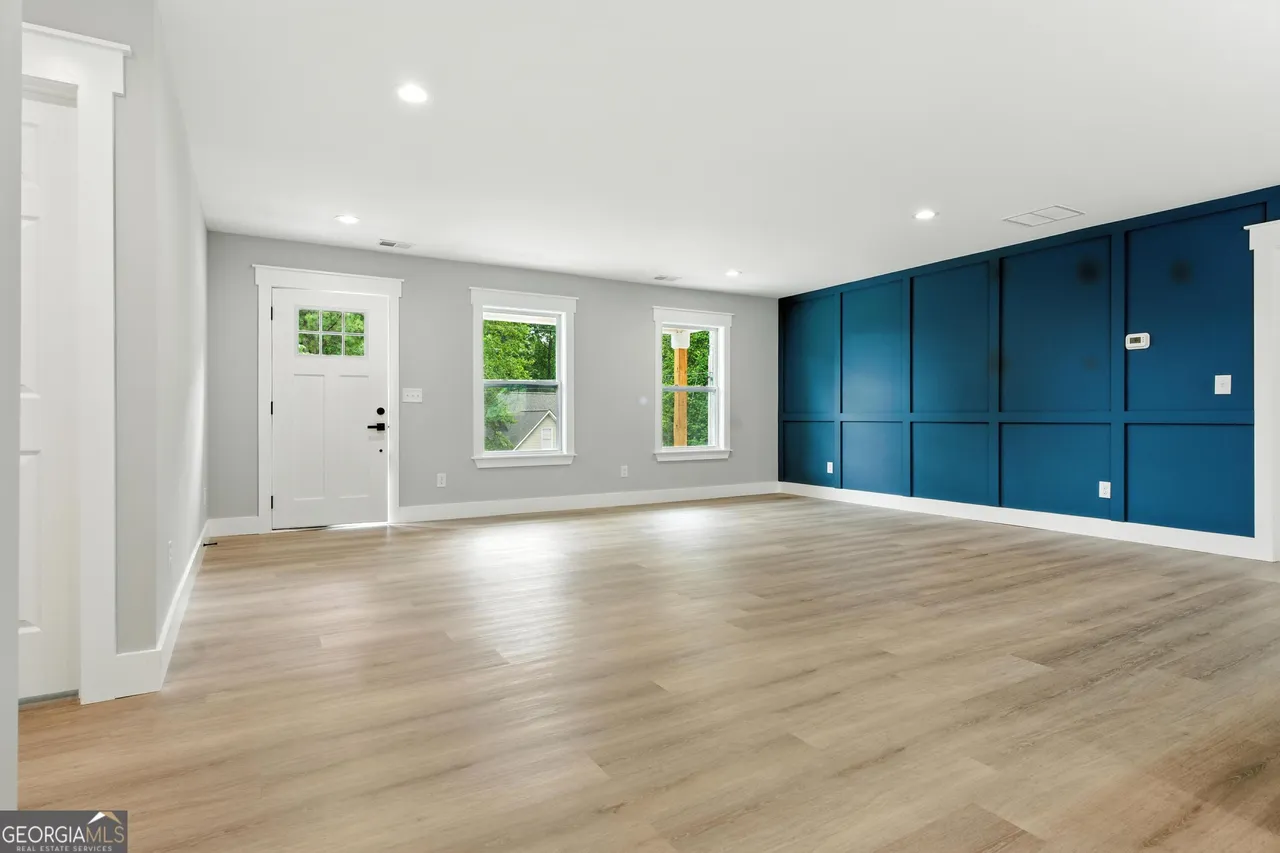 Spacious room featuring light wood flooring, a navy accent wall on the right, three windows, and a white front door. Bright and modern atmosphere.