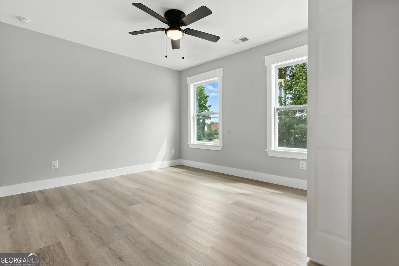A bright, empty room with light wood floors, gray walls, and two large windows. A modern black ceiling fan hangs from the white ceiling, conveying a fresh and airy feel.