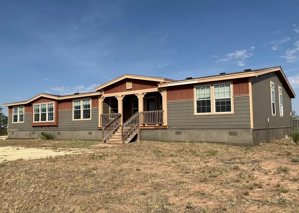 A single-story modular home with a brown and beige exterior stands on dry, sparse grass under a clear blue sky, conveying a serene, rustic atmosphere.