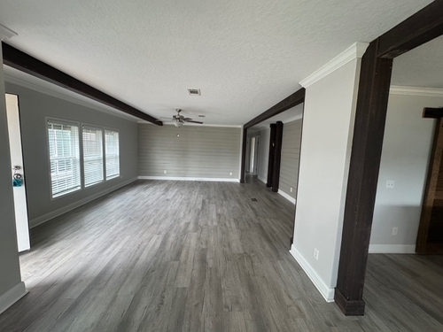 Spacious empty living room with light gray walls, wooden floors, and large windows on the left. Exposed ceiling beams and a ceiling fan add character.