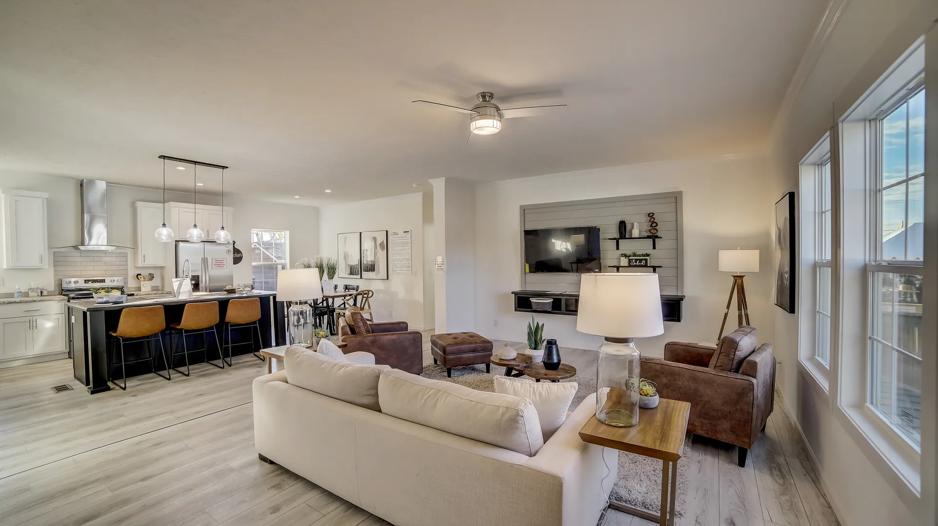Spacious open-concept living room and kitchen with modern decor. White walls, beige sofa, brown armchairs, and a bar with leather stools. Cozy, inviting feel.