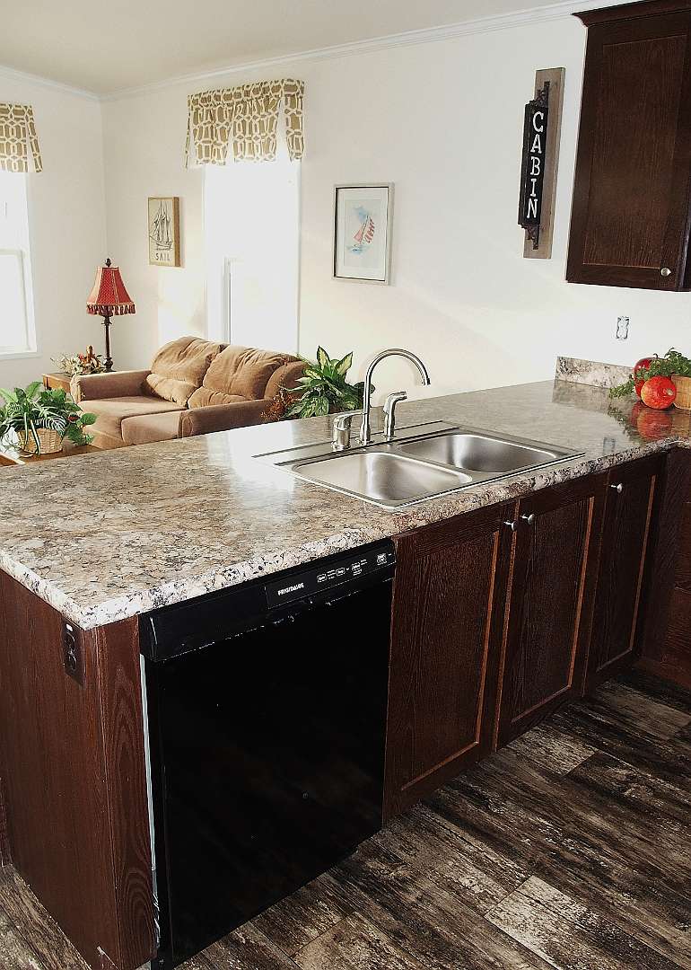 A cozy kitchen space features a marble countertop with a double sink, dark wooden cabinets, and a black dishwasher. A living area with a sofa and lamp is seen in the background, along with wall decor and subtle greenery, creating a warm, inviting atmosphere.