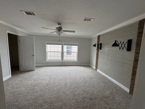 Spacious, empty room with beige carpet, gray wood accent wall, black sconces, ceiling fan, and two large windows, conveying a modern, neutral tone.