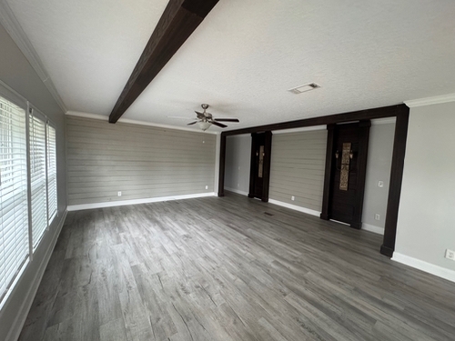 Spacious empty room with light gray walls, large windows on the left, a prominent wood ceiling beam, and a wooden floor, creating a modern, airy feel.