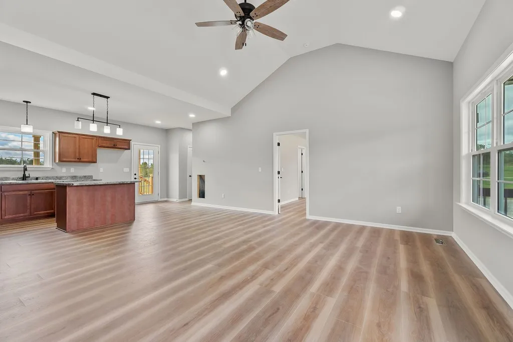 Spacious living area with light wood flooring, gray walls, large window, ceiling fan, and open kitchen with cherry cabinets and granite countertops. Peaceful ambiance.