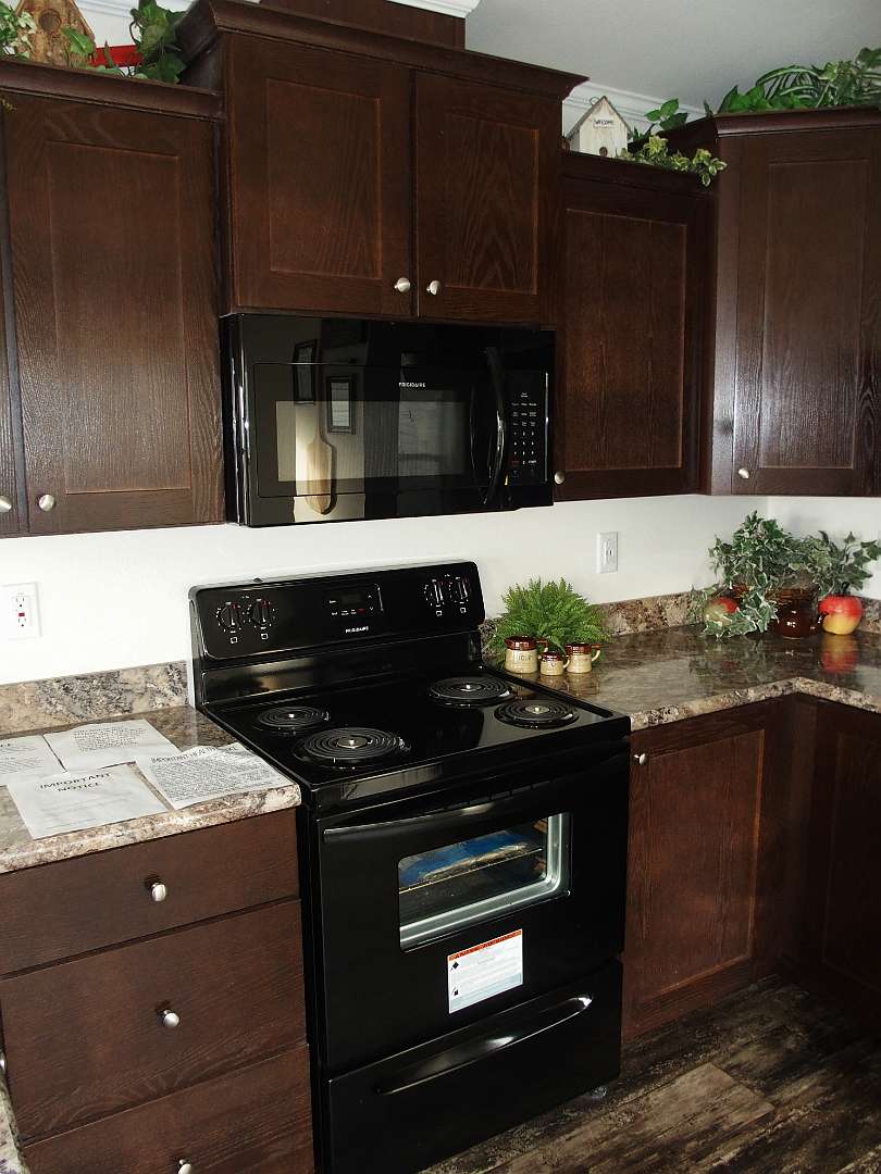 Modern kitchen with dark wooden cabinets and black appliances, including an oven and a microwave. Granite countertop adorned with greenery and fruit. Cozy and elegant atmosphere.