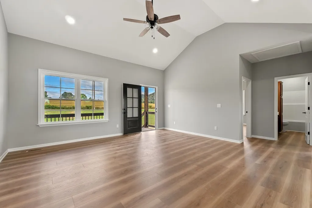 Spacious empty room with light wooden floor, gray walls, a ceiling fan, and large windows. Open door reveals a sunny outdoor view. Minimalist and serene ambiance.