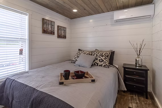 Cozy bedroom with a wooden ceiling and white shiplap walls. The bed has patterned pillows and a tray with mugs. A side table holds branches in a glass vase.