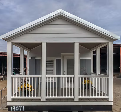 A small, modern gray tiny house with white trim and a gabled roof, features a cozy front porch, stairs, and large windows, set in an outdoor lot.