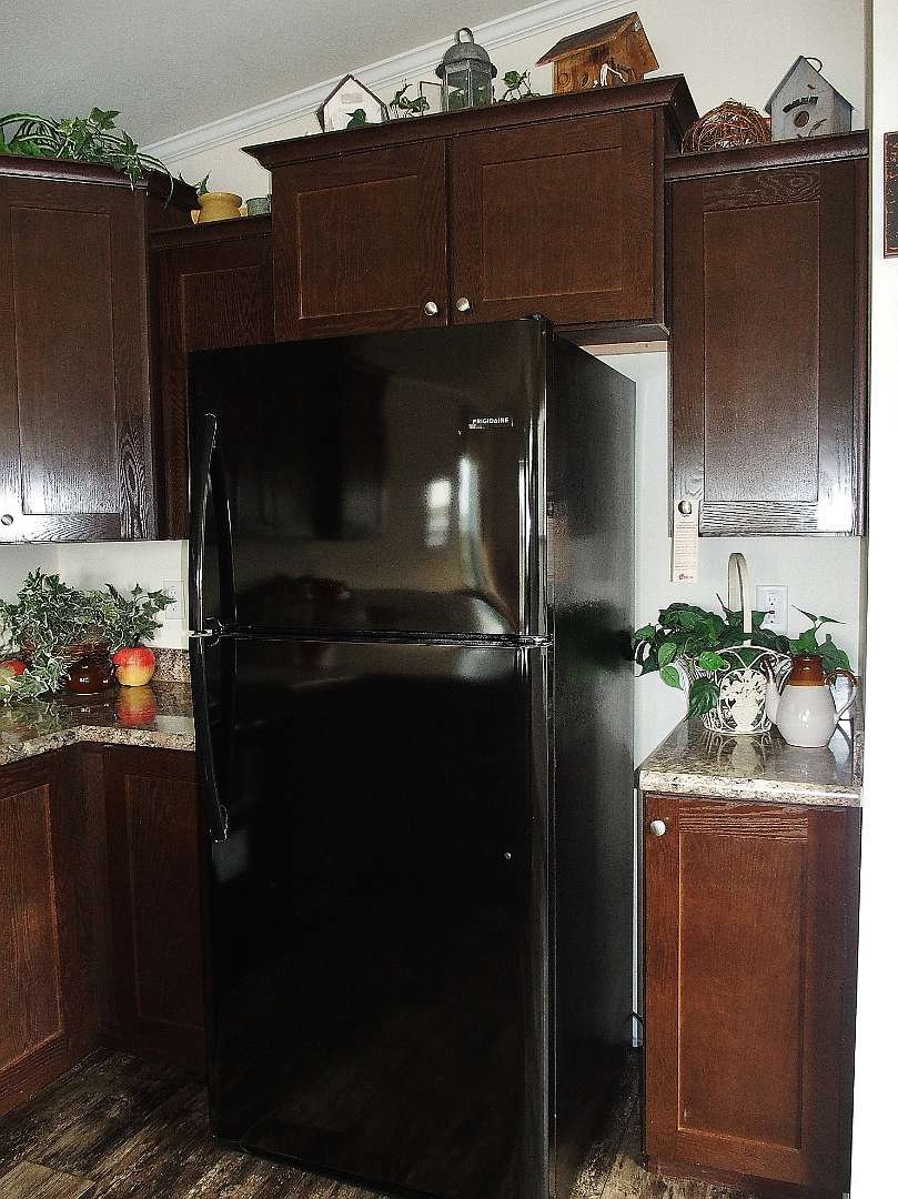 A black refrigerator is surrounded by dark wooden cabinets in a cozy kitchen. Decorative plants and pottery adorn the granite countertops, creating a warm, inviting atmosphere.