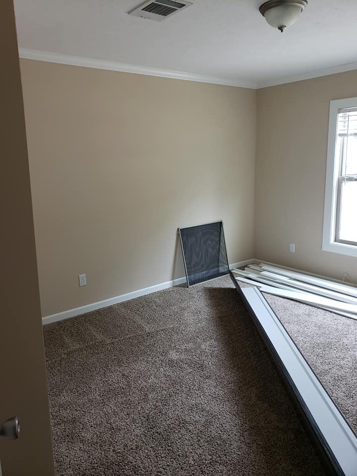 Empty room with beige walls, a window screen leaning against the wall, carpeted floor, and disassembled metal bed frame parts on the floor.