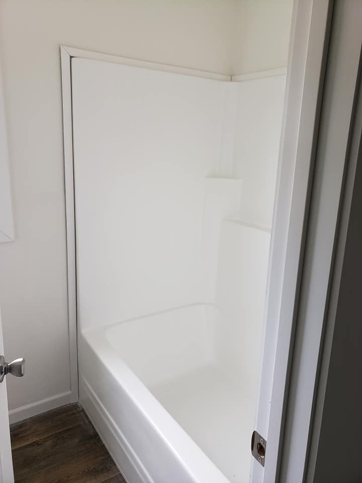 Minimalist bathroom featuring a white bathtub surrounded by plain white walls. The floor is dark wood, creating a clean and modern atmosphere.