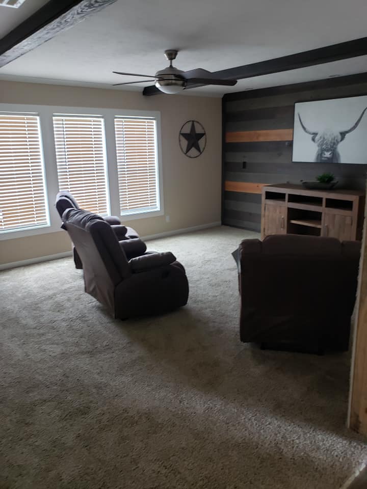 Cozy living room with carpeted floor, featuring two recliners facing a wooden entertainment center. Large windows with blinds and rustic decor add warmth.