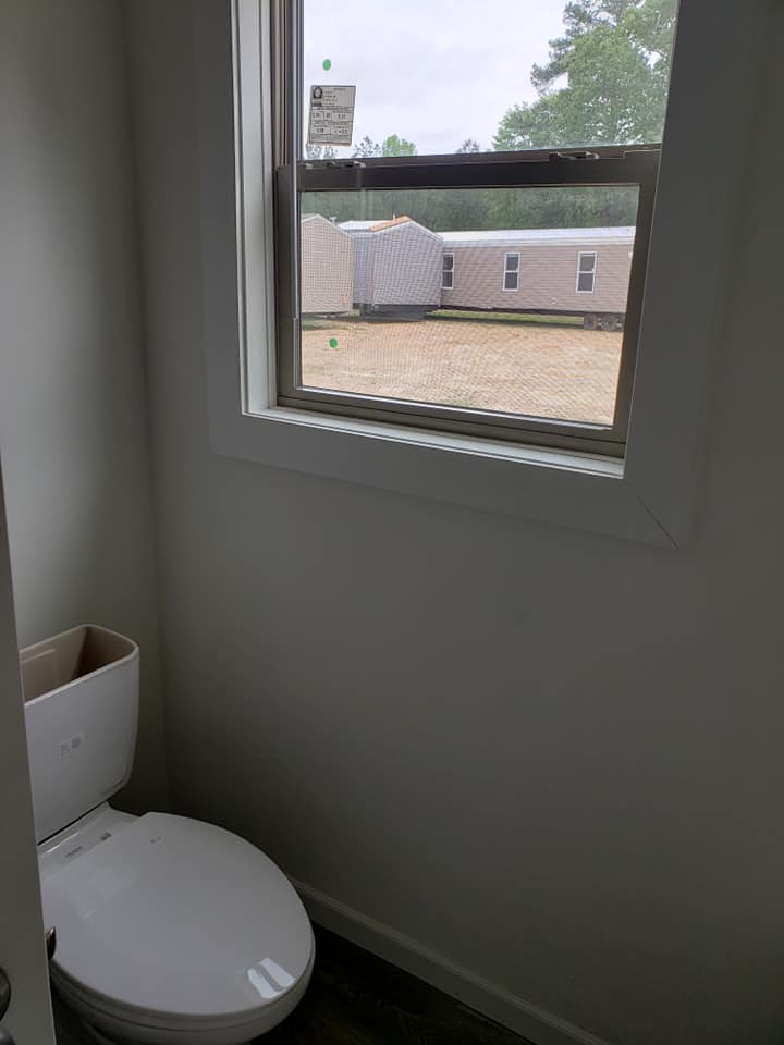 A small bathroom with a white toilet near a window. Outside, mobile homes are visible under a cloudy sky. The setting feels minimal and functional.