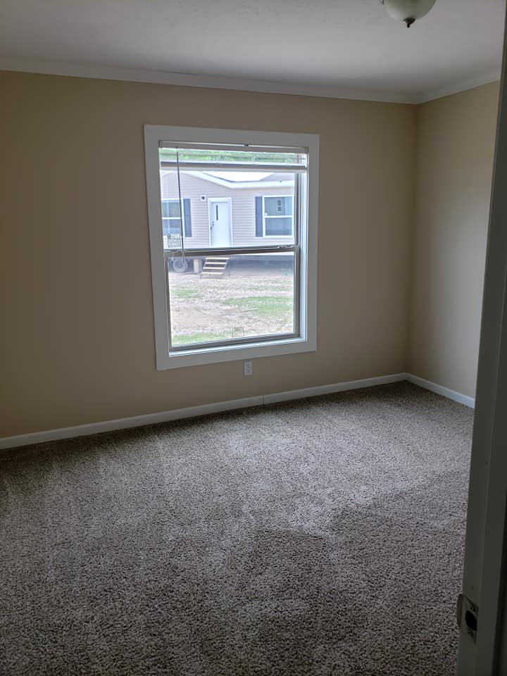 Empty room with beige walls, carpeted flooring, and a single window showing an exterior view of another building. The room feels quiet and bare.