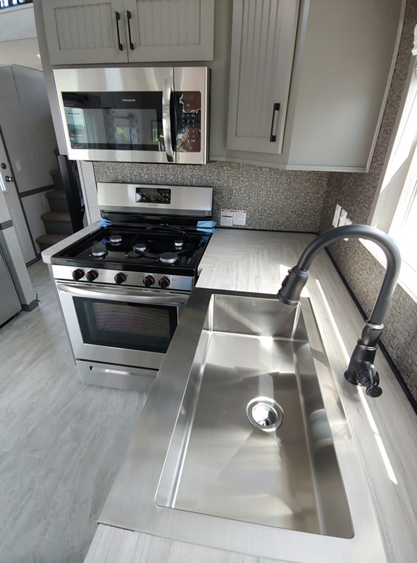 Modern kitchen with sleek stainless steel appliances. Features a large sink, gas stove, overhead microwave, and minimalist cabinets, all bathed in sunlight.