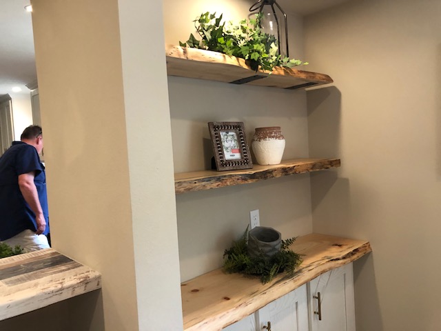 Wooden shelves with rustic decor, including a framed photo, pottery, and plants, against a beige wall. A person in blue is partially visible on the left.