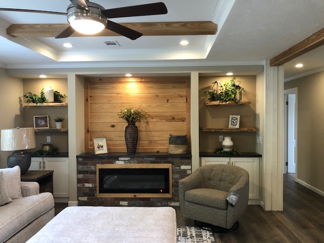 Spacious living room with wood and brick fireplace, cozy gray armchair, ceiling fan, and elegant decor. Warm lighting and neutral tones.