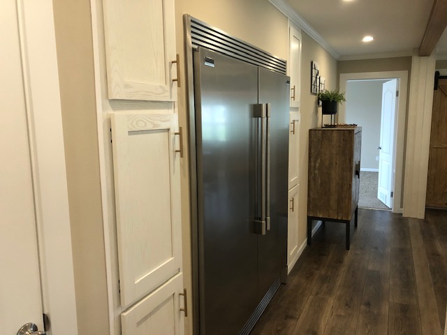 Wide hallway featuring a modern stainless steel refrigerator, surrounded by white cabinets. Wooden flooring and soft lighting create a warm, inviting ambiance.