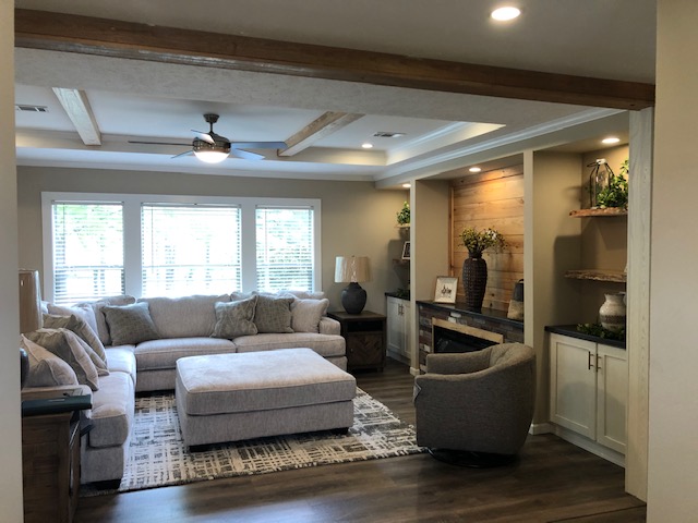 A cozy living room with a large gray sectional sofa and matching ottoman on a patterned rug. Natural light streams through three windows, illuminating the space. A ceiling fan and wooden beams are overhead. Built-in shelves with decor adorn the side wall, creating a warm, inviting ambiance.