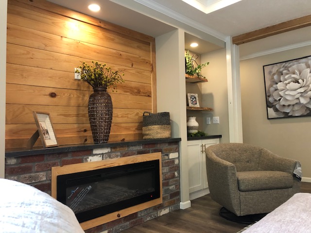 A cozy living room with a modern electric fireplace, wooden accent wall, and built-in shelves. A large vase with flowers and a cushioned chair add warmth.