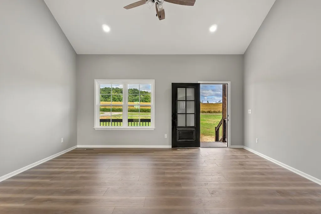 Spacious, empty living room with light gray walls, wooden floor, ceiling fan, large window, and glass-paneled door open to a sunny field.