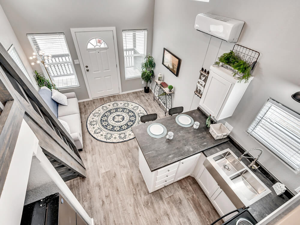Aerial view of a cozy, modern living space with a small kitchen. Features wood flooring, a round patterned rug, and a gray sofa. Bright light filters through two windows, creating an airy feel.