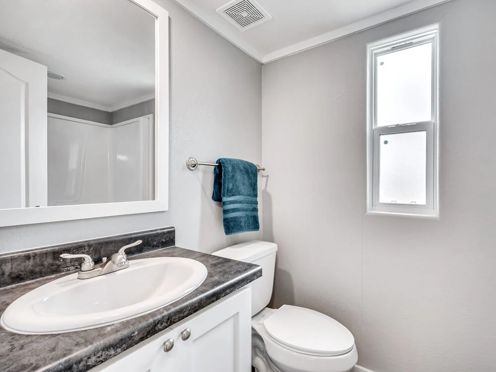 A clean, modern bathroom with gray walls, a white sink atop black countertop, toilet, a small window, and a blue towel on a silver rack.