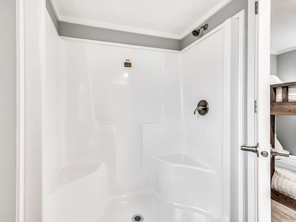 Clean, white shower stall with a chrome faucet and spacious interior in a modern bathroom. Adjacent room features a glimpse of a bunk bed. Neutral tones.