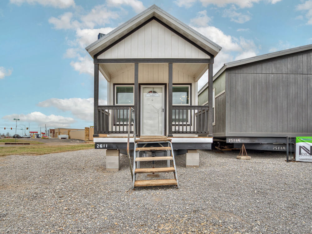 Small, white tiny house with a gabled roof and front porch, elevated on blocks. Wooden stairs lead to the door. Clear blue sky in the background.