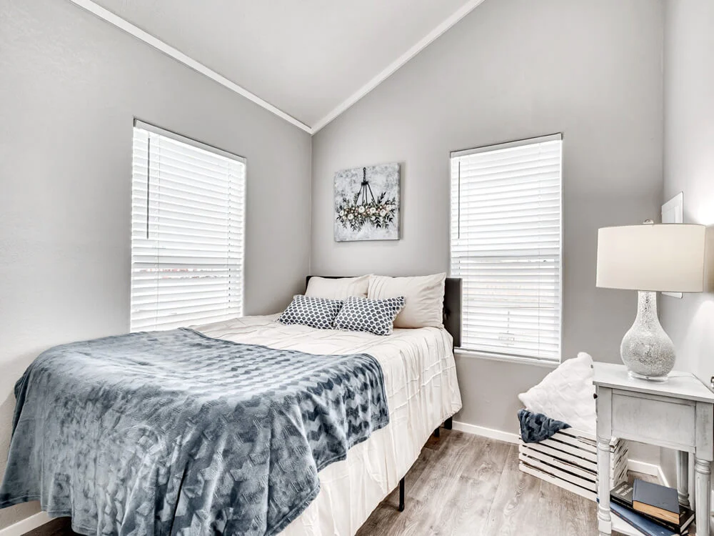 Cozy bedroom with gray walls, featuring a neatly made bed with blue and white bedding, two windows, soft lighting from a table lamp, and floral artwork.