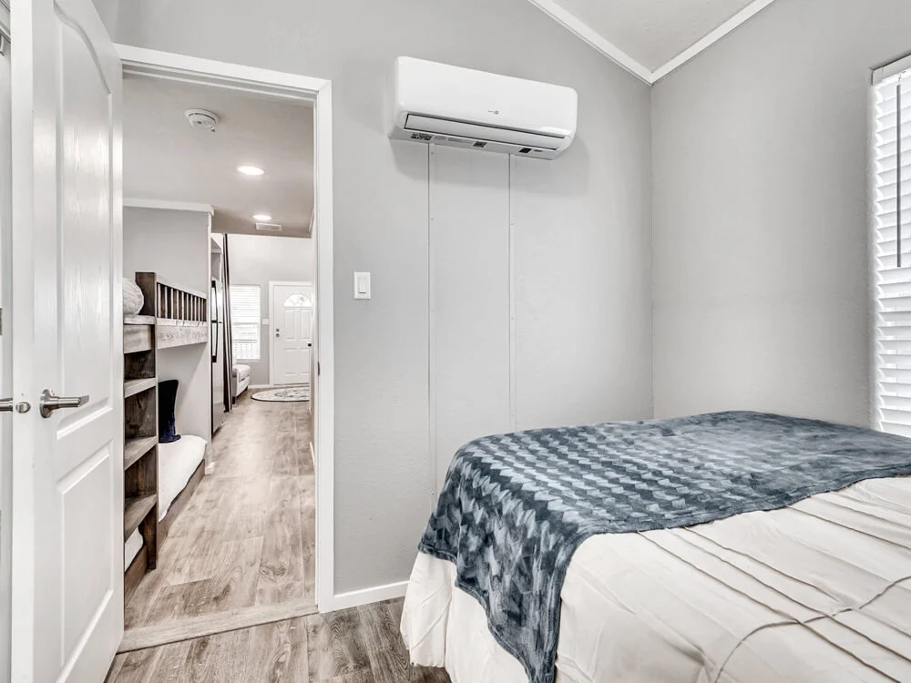 Small bedroom with gray walls and wood flooring, featuring a bed covered in a blue checkered blanket. An open door reveals a hallway with bunk beds and an entrance. An air conditioning unit is mounted on the wall, with natural light filtering through blinds, creating a cozy atmosphere.