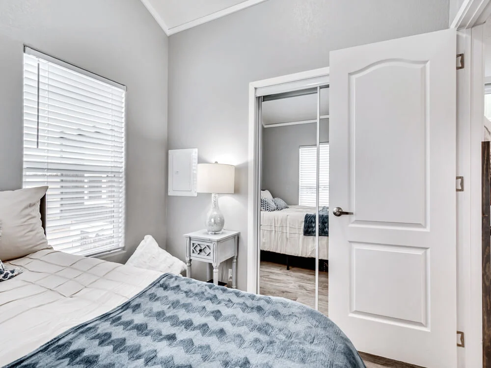 A cozy bedroom with a soft gray color scheme features a bed with white and blue bedding. A bedside table with a lamp sits under a window with blinds, and a mirrored closet door reflects the room. The atmosphere is serene and tidy.